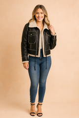 Black faux-leather shearling jacket with front pockets, beige lining, and stitched detailing, styled with slim-fit blue jeans and black heels.